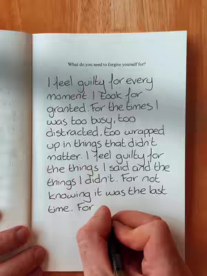 The image shows a close-up of an open book on a wooden surface. Handwriting fills the pages, with one hand visible, in the process of writing on the page. The text focuses on feelings of guilt.