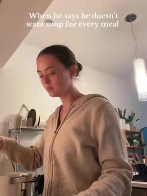 A woman is standing in a kitchen, looking down and to the side with what appears to be a wry expression. She is wearing a beige zip-up hoodie, with a kitchen counter and shelves with dishes and other items in the background. Text overlayed says, "When he says he doesn't want soup for every meal."