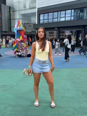 A woman stands confidently outdoors in a sunny setting. She wears a pale yellow tank top and a short denim skirt. Her hair is long and dark, and she has a neutral expression. The background shows a mix of buildings and people, and in the distance stands a colorful Pride display.