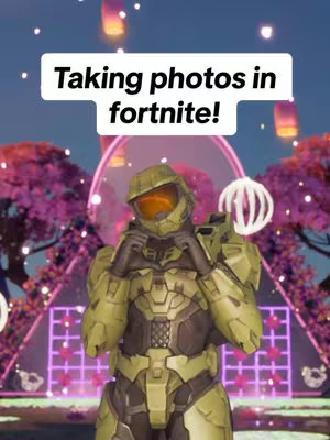 The image features a character from the game Fortnite dressed in green armor, resembling Master Chief from Halo. The character is posed in front of a backdrop with pink flowering trees, paper lanterns, and a circular light. White text in a rectangular box at the top reads 'Taking photos in fortnite!'