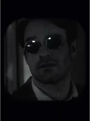 A grayscale image featuring a man with dark sunglasses and a serious expression. The composition focuses on his face. The lighting is dim, and the man's attire suggests a formal setting.