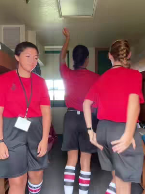 The cover image features three individuals, likely cadets, standing in what appears to be a military school setting. The composition is centered on the three figures, all wearing red shirts and dark shorts, with matching striped socks. One is raising their arm, while another is turned in the opposite direction. The lighting seems natural, possibly indoors, with no prominent background details.