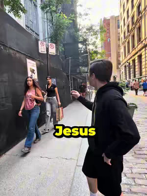 A man in a black hoodie gestures towards three people walking on a city street. The word 'Jesus' is highlighted in a yellow box on the lower left.