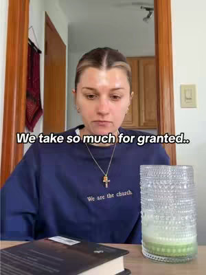 A woman sits at a table, looking down with a thoughtful expression. She is wearing a dark blue sweatshirt with text on it and a gold cross necklace. In the foreground is an open book and a glass candle holder, which reflects light. The background shows a glimpse of a kitchen doorway.