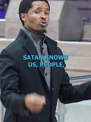 A medium shot shows a Black man in a black overcoat and gray turtleneck, speaking directly to the camera. Text that reads "SATAN KNOWS US, PEOPLE," is superimposed over the front of his coat. The background is blurred, with what appears to be a stairway and a gray architectural feature.