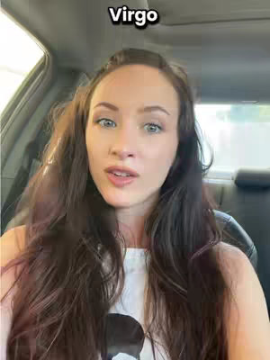 A medium shot taken from inside of a car featuring a woman with long, dark brown hair facing the camera. She is wearing a white t-shirt. Above her head, a white rectangular text box reads "Virgo". The interior of the car, including the headrest and a portion of the window, frames the shot.