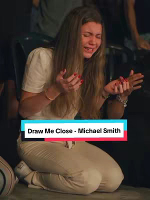 A woman kneels, hands raised in supplication, with a tear-streaked face. She is in a performance setting, with others in the background, a text overlay is at the bottom of the image.
