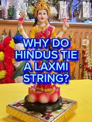 The image features a vibrant depiction of a Hindu deity statue, likely Lakshmi, amidst a colorful display of worship items. The central focus is on the statue of a female deity, adorned with a crown and jewelry, and holding items in her hands. Bold, blue text overlaid on the image poses a question about the significance of tying a Lakshmi string. The overall composition is a colorful, eye-catching, and inviting visual with elements of religious devotion.