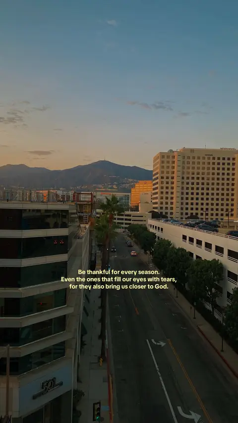 An elevated shot captures a city street scene at dusk. The sky transitions from orange to blue, with buildings and mountains making up the skyline. The foreground features a tree-lined road with cars, and text overlays. The overall tone is peaceful and inspirational.