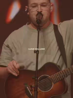 A man with a shaved head and glasses sings into a microphone, holding a guitar. The background has blurred stage lights. The words 'I LOVE YOU, LORD' are overlaid on the image.