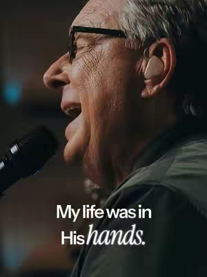 A medium shot spotlights a man with gray hair and glasses singing into a microphone. He has a serious expression, and text overlays the image.