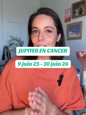 A woman with dark hair poses for the camera, her upper body visible against an interior backdrop. She wears an orange t-shirt and a small black microphone is clipped to it. Overlayed on the image are two white rectangular boxes containing the text 

JUPITER EN CANCER

and

9 juin 25 - 30 juin 26

The background is softly blurred, featuring parts of a door, framed pictures, and a few decorative items. The lighting appears natural.
