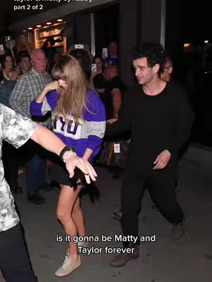 A paparazzi photo captures Taylor Swift and Matty Healy navigating a crowd outside a building. Taylor is smiling, and appears to be acknowledging the camera. They are surrounded by onlookers who are eagerly trying to get a glimpse of them and take photos.