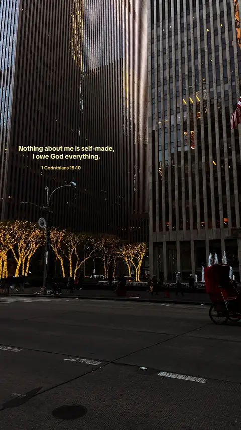 The image captures a street scene at dusk or night, with towering skyscrapers dominating the background. The buildings are made of a dark, reflective material, creating a sense of verticality. In the foreground, a street is visible, with a row of trees lit up with yellow lights. The overall lighting is dim, casting shadows and giving the scene a cool, urban atmosphere.