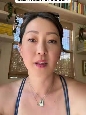 A woman with dark hair and sunglasses perched on her head is centered in the frame, with a well-lit background. She is wearing a blue tank top and a necklace. Behind her is a window with blinds, and a shelf filled with books and a few plants.