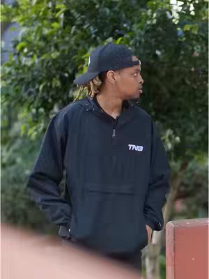 A man wearing a black cap and jacket with a white logo, standing in front of green foliage. The focus is on him, with the background slightly blurred. The composition presents a casual outdoor scene.