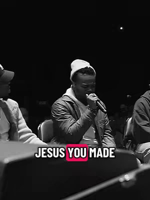 A black and white image with three people on stage, the center person singing with a microphone while wearing a beanie and jacket. Text overlayed at the bottom reads, "JESUS YOU MADE."