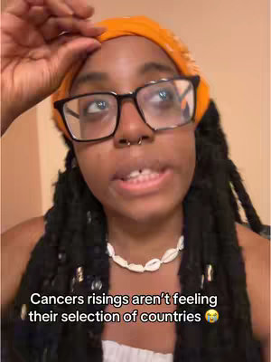 A close-up shot of a young woman with black hair and glasses. She wears an orange bandanna, a white necklace, and has a septum piercing. Text overlayed at the bottom of the image reads, "Cancers risings aren't feeling their selection of countries ๐ฅบ."