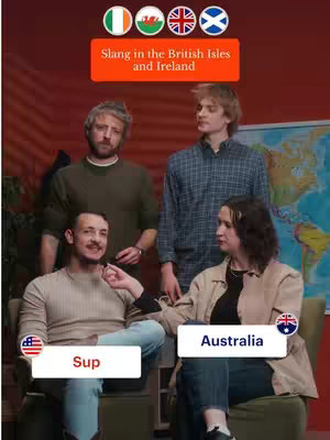 The image is a bright, studio-lit shot featuring four individuals of diverse ethnicities, positioned in front of a maroon backdrop and a partially visible world map. The foreground includes two figures, a man on the left and a woman on the right, seated. The man in the foreground, who's focused on the left talking, is dressed in a light-colored sweater over blue jeans. He is holding a small item, with a word bubble from the US flag that says "Sup". The woman is seated on a chair to the right wearing a beige jacket and blue jeans. Each of them is in close dialogue, interacting with each other. Beside the woman is a word bubble with the Australian flag and the word "Australia". They are facing one another, as if in conversation, two additional people standing behind them. On the top of the image there are four flags: Irish, Welsh, Great Britain, Scottish.