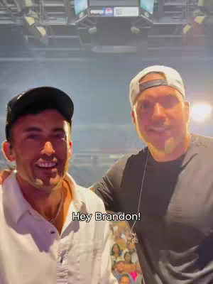 Two men are smiling in a selfie taken inside a large venue. The man on the left is wearing a white shirt and a black cap. The man on the right is wearing a black shirt and a white cap.