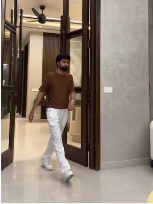 A man in a brown t-shirt and white pants is walking through a doorway. The background is a well-lit hallway with wooden doors, white walls, and a ceiling fan. The overall composition is clean and modern.