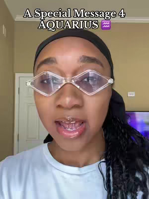 A person with dark skin, wearing clear, diamond-shaped glasses, and a black head covering, is the focal point. The person is looking directly at the camera with their mouth slightly open. Overlaid on the image, at the top, is text that reads, "A Special Message 4 AQUARIUS" with an Aquarius symbol. The background is a neutral beige, with a light-colored door frame on the left and a hint of a television screen visible on the right.