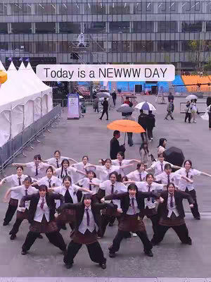 The image shows a group of young women, dressed in school uniforms with white shirts, dark blazers, and skirts, posing with their arms outstretched in a dance position in front of a modern glass building. People with umbrellas and walking around, indicating an outdoor setting. A white banner with the text 'Today is a NEWWW DAY' is hovering above them. 