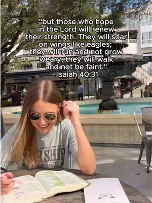 A young woman with blonde hair and sunglasses sits at a table outdoors, leafing through a book. The background shows a small fountain and buildings.