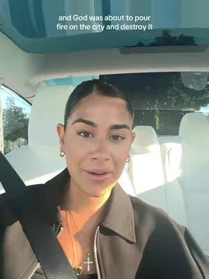A woman is seated in a car, facing forward and speaking to the camera. She has dark hair and is wearing earrings and a necklace with a cross pendant. She wears a brown jacket. The car's windshield and a portion of the car's interior are visible in the background.