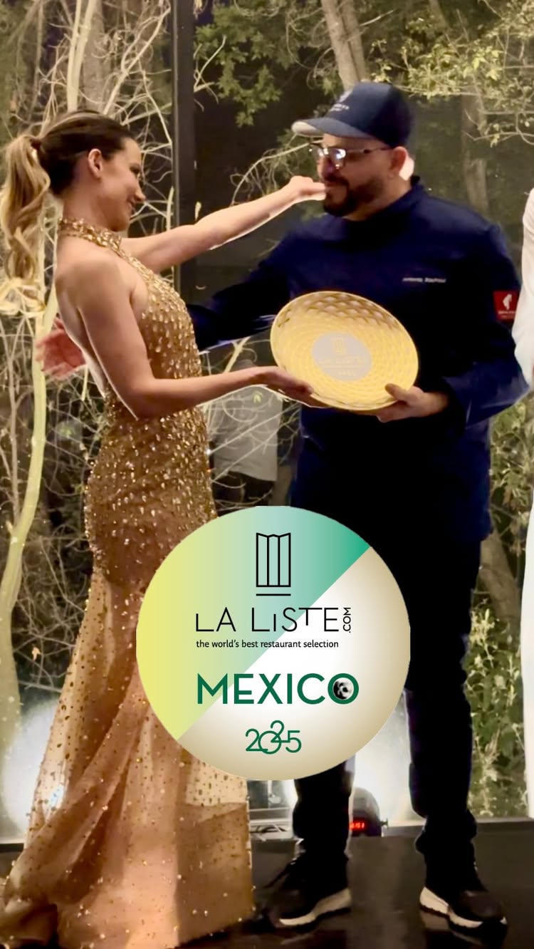 The image shows a woman in a gold dress receiving a trophy from a man in a dark blue suit. A logo with the words 'LA LISTE' and 'MEXICO' is below the people. They are in front of a blurred backdrop of trees and lights.