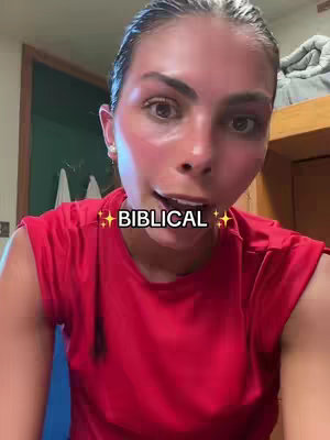 A close-up shot of a woman with fair skin and dark hair, appearing red-faced as if she has been exerting herself. She has visible pink blush over her cheeks and temples. She is facing the camera and talking with a look of concentration on her face and has text overlayed with an overlay that says 'BIBLICAL'. She wears a red t-shirt. The background is slightly blurred, featuring a doorway and some interior decor suggestive of a home or dormitory.