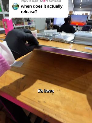 A shot from inside a workspace, featuring a wooden desk with a reflective metal strip. A hand in a black glove is working on the desk. In the background, a computer screen displays a sunset scene.