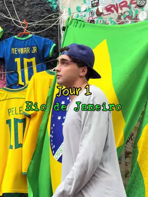 A young man stands in front of a partially visible wall and a large Brazilian flag. He is wearing a white shirt and a dark blue cap. Behind him, there are football jerseys hanging. The words "Jour 1 Rio de Janeiro" are written on the image.