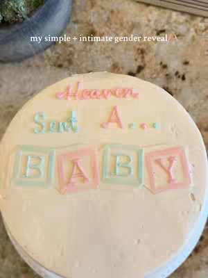 A round cake with white frosting. In the middle, it has letters in pink and green frosting that say: Heaven Sent A... with the word 'BABY' spelled out in colored blocks below.