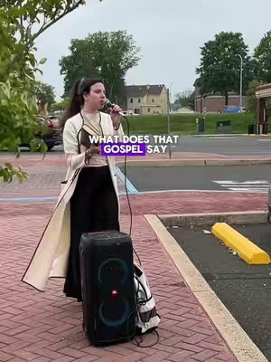 A woman stands outdoors holding a microphone, speaking into it with what appears to be a speaker behind her. She is wearing a white coat and black pants and is standing in front of a gray building.