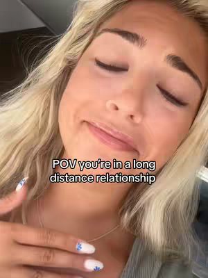 The close-up shot features a young woman with blonde hair, gazing slightly away from the camera with a mix of emotions on her face. Her eyes are partially closed, and she has a subtle, almost melancholic smile. The text overlay states "POV you're in a long distance relationship". Her hand, adorned with nail art featuring flower designs, is visible in the frame. The background is blurred, keeping the focus on the woman's face and expression.
