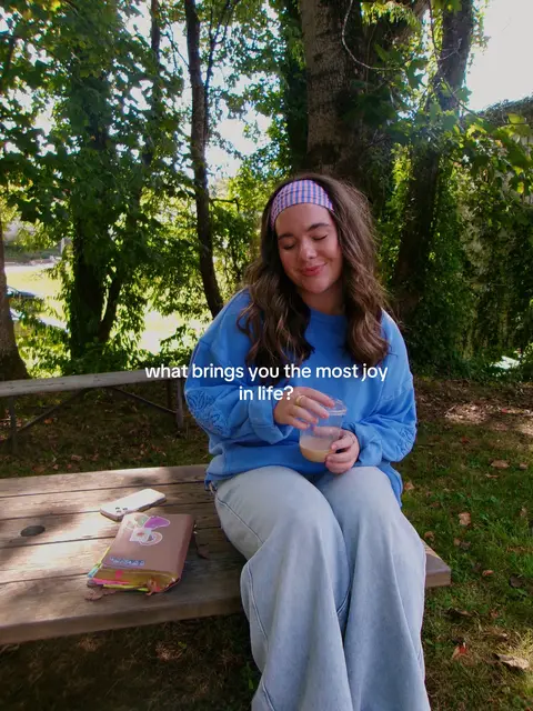 A young woman with long hair and a headband sits on a wooden picnic table in a park-like setting. She is holding a clear cup with a light brown liquid and smiling slightly with her eyes closed. The background includes green trees and foliage, adding to the natural, serene atmosphere. A smartphone and a book are on the table beside her.
