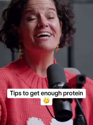 A woman with curly dark hair, wearing a red sweater, and holding a microphone, is speaking. A text overlay at the bottom says "Tips to get enough protein" with a thinking face emoji. The background is blurred.
