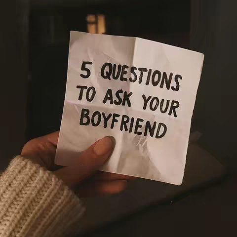 A hand holds a crumpled white piece of paper against a dark background. The paper has the text "5 QUESTIONS TO ASK YOUR BOYFRIEND" written on it in black, handwritten-style letters. The hand is fair-skinned, with visible skin and a light-colored sweater sleeve.
