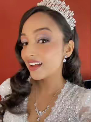 A medium shot of a woman in profile smiling with an open mouth, eyes looking slightly upwards. She's wearing a tiara, earrings, and a necklace, with a red background. Her dark hair is styled, and she's wearing makeup.