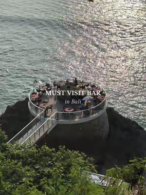An aerial shot captures a bar built on the edge of a cliff, overlooking the ocean. The bar has a circular design with tables and seating under a roof. The text overlay reads 'MUST VISIT BAR in Bali'. The water shimmers in the sunlight, and the greenery of the cliff frames the scene.