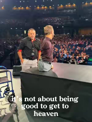 The image shows a live event with a man speaking on a stage. Behind him, a large crowd is visible. The foreground includes a dark stage element and a wheelchair.