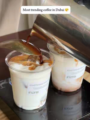 The image shows a close-up of two clear plastic cups filled with an iced beverage. Coffee is being poured from a stainless steel pitcher into one of the cups. The beverage appears to be layered with a light, creamy base, ice cubes, and a darker liquid on top. Text overlays indicate the drink is an iced breve macchiato from FLTR in Dubai.