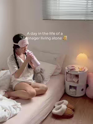 A woman in pajamas is seated on a bed. She has a sleep mask on and is drinking from a water bottle. The bedroom has a window, a bedside table, and a lamp.