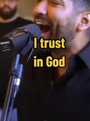 A man singing into a microphone with the text overlay "I trust in God". The image is close-up focused on the man's face and the microphone. The background appears to be a stage or studio setting, with another person partially visible. The color palette is warm, dominated by skin and dark tones, with the text emphasized in yellow.
