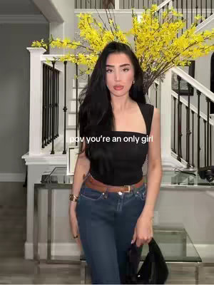 A woman stands in a well-lit entryway, wearing a black top and jeans, with text overlaying the image. Behind her is a staircase and large floral arrangement.