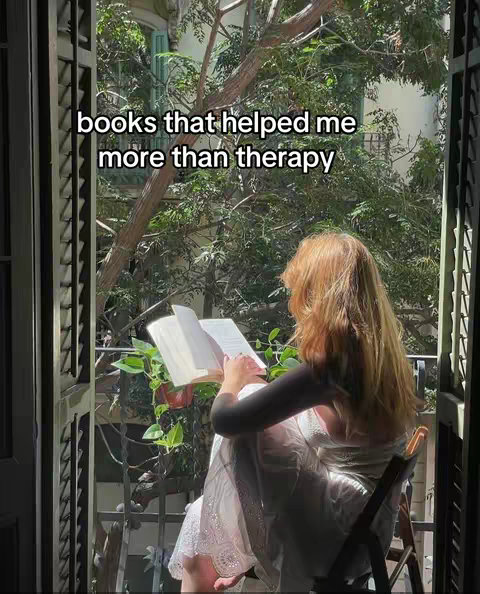A woman with long blonde hair sits on a chair on a balcony reading a book. The scene is bathed in sunlight, with green trees and foliage visible through the balcony's railings. The woman wears a white dress and the overall aesthetic is serene and inviting.
