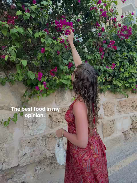 A woman in a red and gold patterned dress stands outdoors near a stone wall. She is reaching up towards a bush of pink flowers. The scene is brightly lit.