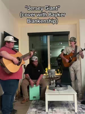 The cover image shows three men playing music in a living room. The man on the left is playing an acoustic guitar and wearing a white hat and pink shirt. In the middle, a man sits playing a cajon drum and wearing a black shirt and glasses. The man on the right is also playing an acoustic guitar and wearing a hat and light-colored clothes. The text overlay says "Jersey Giant" (cover with Bayker Blankenship) in a white, bold font. The setting includes living room furniture against a neutral background.