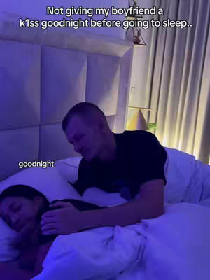 A man is leaning over a sleeping woman in a bed. Above, text reads, "Not giving my boyfriend a kiss goodnight before going to sleep." The man is wearing a black shirt, and the lighting is dim and blue-toned. The scene is framed to include part of the headboard and surrounding bedroom.